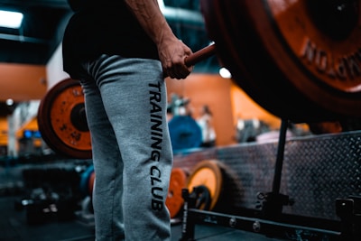 A person wearing grey sweatpants with the words 'TRAINING CLUB' printed on the side is holding a barbell in a gym setting. The barbell is loaded with large, orange weight plates. The background shows gym equipment and has a blurred effect, indicating a focus on the person and the barbell. The environment is industrial, with metallic surfaces and equipment visible.