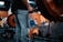 A person wearing grey sweatpants with the words 'TRAINING CLUB' printed on the side is holding a barbell in a gym setting. The barbell is loaded with large, orange weight plates. The background shows gym equipment and has a blurred effect, indicating a focus on the person and the barbell. The environment is industrial, with metallic surfaces and equipment visible.