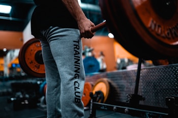 A person wearing grey sweatpants with the words 'TRAINING CLUB' printed on the side is holding a barbell in a gym setting. The barbell is loaded with large, orange weight plates. The background shows gym equipment and has a blurred effect, indicating a focus on the person and the barbell. The environment is industrial, with metallic surfaces and equipment visible.