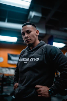 A man wearing a dark hoodie printed with 'Training Club RFW.' stands confidently in a gym setting. The background is softly blurred, highlighting a few wall decorations and warm lighting.