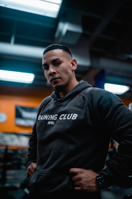 A man wearing a dark hoodie printed with 'Training Club RFW.' stands confidently in a gym setting. The background is softly blurred, highlighting a few wall decorations and warm lighting.