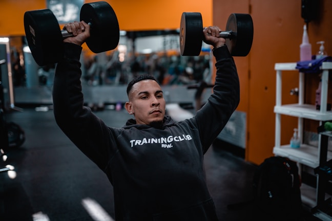 A person is lifting two large dumbbells overhead while wearing a dark hoodie that reads 'Training Club.' The setting appears to be a gym with blurred gym equipment in the background and orange walls.
