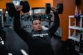 A person is lifting two large dumbbells overhead while wearing a dark hoodie that reads 'Training Club.' The setting appears to be a gym with blurred gym equipment in the background and orange walls.