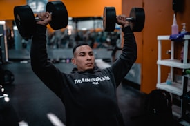 A person is lifting two large dumbbells overhead while wearing a dark hoodie that reads 'Training Club.' The setting appears to be a gym with blurred gym equipment in the background and orange walls.