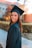 A young woman wearing a graduation cap and gown, smiling while standing outdoors. The background shows a building and trees with autumn foliage, suggesting a college or university campus.