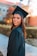 A young woman wearing a graduation cap and gown, smiling while standing outdoors. The background shows a building and trees with autumn foliage, suggesting a college or university campus.