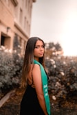 A young woman with long dark hair stands outdoors, wearing a black dress and a green sash with the words 'Class of 2021'. The background consists of a blurred garden with some bushes and a building.