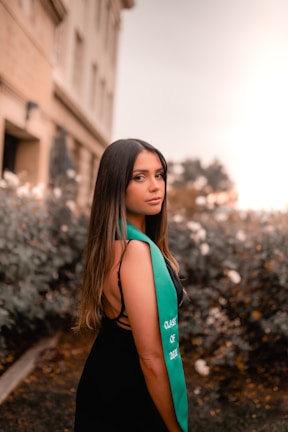 A young woman with long dark hair stands outdoors, wearing a black dress and a green sash with the words 'Class of 2021'. The background consists of a blurred garden with some bushes and a building.