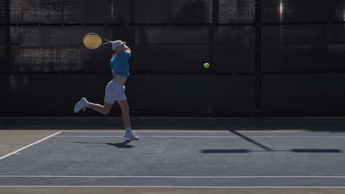 A tennis player is captured mid-action, performing a backhand stroke on a tennis court. The player is wearing a blue shirt and white shorts, with a cap on their head. The tennis ball is in mid-air just before impact with the racket. The court surface appears dark, contrasting with the bright colors of the player’s outfit and the ball.