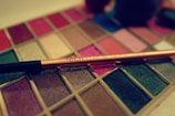 An assortment of vibrant makeup colors is displayed in a palette. A shiny, gold-toned makeup brush labeled 'Real Techniques' is placed diagonally across the palette. The palette features a variety of shades, including reds, pinks, browns, blues, and greens, some with a glittery finish.