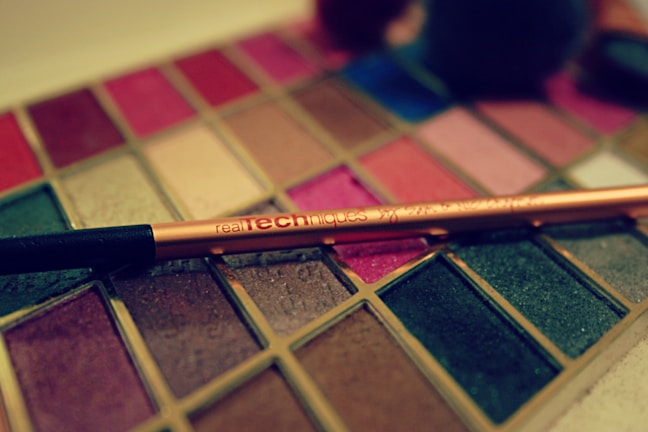 An assortment of vibrant makeup colors is displayed in a palette. A shiny, gold-toned makeup brush labeled 'Real Techniques' is placed diagonally across the palette. The palette features a variety of shades, including reds, pinks, browns, blues, and greens, some with a glittery finish.