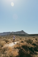A person is running on a narrow dirt trail through a dry, rugged landscape with sparse vegetation. In the background, there are flat-topped mountains under a clear blue sky with the sun shining brightly.