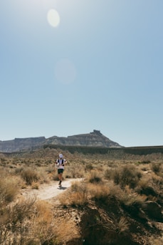 A person is running on a narrow dirt trail through a dry, rugged landscape with sparse vegetation. In the background, there are flat-topped mountains under a clear blue sky with the sun shining brightly.