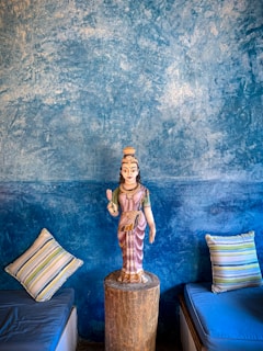 A detailed wall painting with rich textures in shades of blue serves as a backdrop for a statue. The statue is of a traditional figure placed on a wooden pedestal. On either side, striped cushions rest on what appear to be low seating areas.