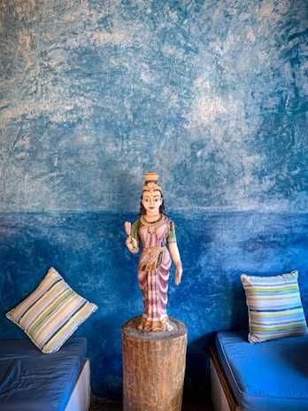 A detailed wall painting with rich textures in shades of blue serves as a backdrop for a statue. The statue is of a traditional figure placed on a wooden pedestal. On either side, striped cushions rest on what appear to be low seating areas.