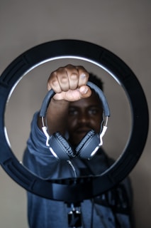 A person holding a pair of black headphones extended towards the camera, framed within a circular ring light. The background is neutral, and the lighting highlights the headphones sharply.