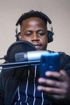 A person wearing headphones is seated in front of a microphone, appearing to be recording or speaking. They are holding a smartphone in one hand.