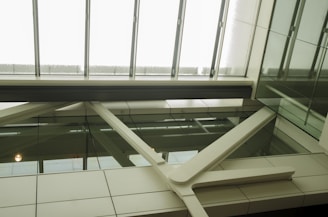 A modern architectural interior view with glass panels and metal beams. The ceiling is composed of large glass skylights, allowing natural light to illuminate the space. The structure features clean lines with a mix of clear and opaque surfaces, creating a sleek and contemporary aesthetic.