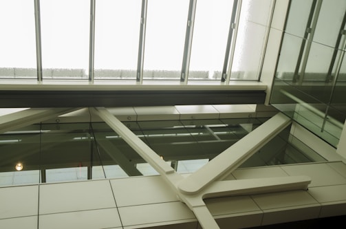 A modern architectural interior view with glass panels and metal beams. The ceiling is composed of large glass skylights, allowing natural light to illuminate the space. The structure features clean lines with a mix of clear and opaque surfaces, creating a sleek and contemporary aesthetic.