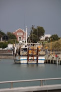 A fishing boat docked at a harbor with a church and several buildings in the background. The boat is loaded with equipment, and there are trees and greenery around the structures.