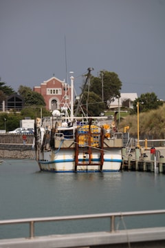 A fishing boat docked at a harbor with a church and several buildings in the background. The boat is loaded with equipment, and there are trees and greenery around the structures.