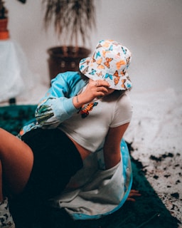 A person is lounging indoors, wearing a white bucket hat adorned with colorful butterfly patterns and a matching jacket. They are dressed in a white crop top and black shorts, sitting on a green textured surface. The setting includes some plants in the background, contributing to a relaxed and artistic atmosphere.