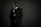 A person in a dark suit, wearing sunglasses and a security badge, stands against a geometric-patterned background. The lighting is low, creating a moody atmosphere.