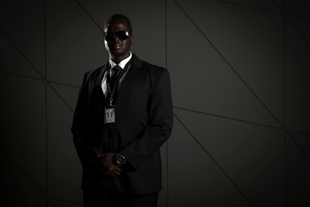 A person in a dark suit, wearing sunglasses and a security badge, stands against a geometric-patterned background. The lighting is low, creating a moody atmosphere.