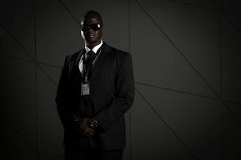 A person in a dark suit, wearing sunglasses and a security badge, stands against a geometric-patterned background. The lighting is low, creating a moody atmosphere.