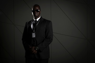 A person in a dark suit, wearing sunglasses and a security badge, stands against a geometric-patterned background. The lighting is low, creating a moody atmosphere.
