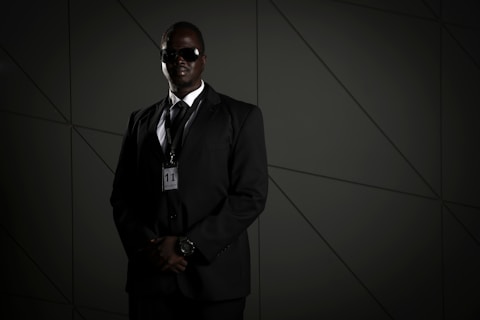 A person in a dark suit, wearing sunglasses and a security badge, stands against a geometric-patterned background. The lighting is low, creating a moody atmosphere.