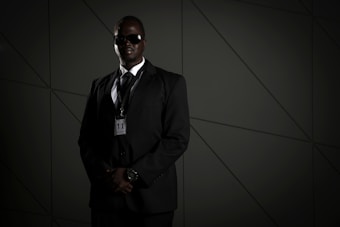 A person in a dark suit, wearing sunglasses and a security badge, stands against a geometric-patterned background. The lighting is low, creating a moody atmosphere.
