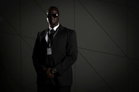 A person in a dark suit, wearing sunglasses and a security badge, stands against a geometric-patterned background. The lighting is low, creating a moody atmosphere.