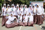 A group of students in white hijabs and plaid skirts are posing together outdoors. Some are holding up their hands, which are painted in vibrant colors like red, green, and yellow. They are smiling and appear to be enjoying a school event. In the background, there is a banner with text, and the surroundings are bright and sunny with trees and school buildings.