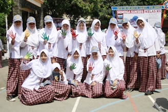 A group of students in white hijabs and plaid skirts are posing together outdoors. Some are holding up their hands, which are painted in vibrant colors like red, green, and yellow. They are smiling and appear to be enjoying a school event. In the background, there is a banner with text, and the surroundings are bright and sunny with trees and school buildings.