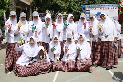 A group of students in white hijabs and plaid skirts are posing together outdoors. Some are holding up their hands, which are painted in vibrant colors like red, green, and yellow. They are smiling and appear to be enjoying a school event. In the background, there is a banner with text, and the surroundings are bright and sunny with trees and school buildings.