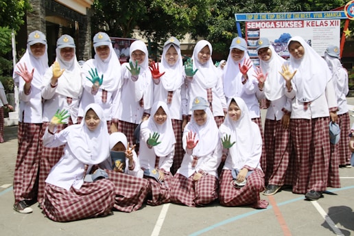 A group of students in white hijabs and plaid skirts are posing together outdoors. Some are holding up their hands, which are painted in vibrant colors like red, green, and yellow. They are smiling and appear to be enjoying a school event. In the background, there is a banner with text, and the surroundings are bright and sunny with trees and school buildings.