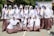 A group of students in white hijabs and plaid skirts are posing together outdoors. Some are holding up their hands, which are painted in vibrant colors like red, green, and yellow. They are smiling and appear to be enjoying a school event. In the background, there is a banner with text, and the surroundings are bright and sunny with trees and school buildings.
