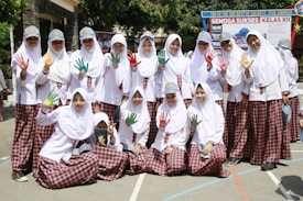 A group of students in white hijabs and plaid skirts are posing together outdoors. Some are holding up their hands, which are painted in vibrant colors like red, green, and yellow. They are smiling and appear to be enjoying a school event. In the background, there is a banner with text, and the surroundings are bright and sunny with trees and school buildings.