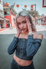 A person with long blonde hair is posing outdoors in front of traditional Asian architecture. They are wearing a denim off-the-shoulder top and matching jeans. Red lanterns are hung around the building, giving a festive look.
