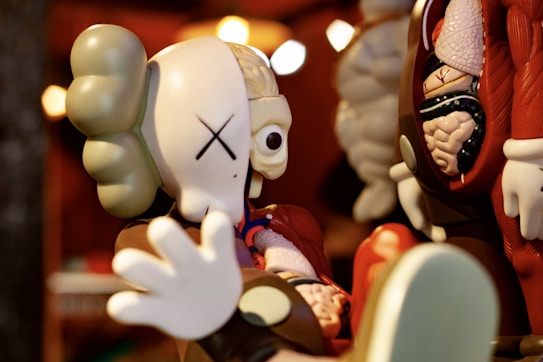 A stylized toy figure with a large head and an anatomical cutaway revealing internal organs. The figure has exaggerated features including large hands and a simplified facial expression with eyes represented by crosses. The toy is reminiscent of a character with a cartoonish anatomy, juxtaposing playful design with anatomical detail.