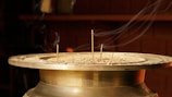 A brass incense holder is filled with ash, from which several incense sticks protrude. Delicate streams of smoke trail upwards, highlighted against a dark, blurred background, suggesting a serene and contemplative atmosphere.