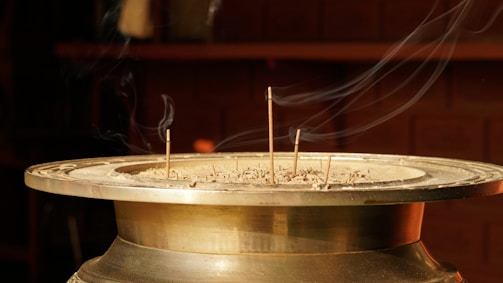 A brass incense holder is filled with ash, from which several incense sticks protrude. Delicate streams of smoke trail upwards, highlighted against a dark, blurred background, suggesting a serene and contemplative atmosphere.