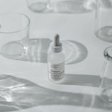 A minimalist arrangement featuring a dropper bottle of skincare product, surrounded by clear, empty glass containers casting soft shadows on a white surface.