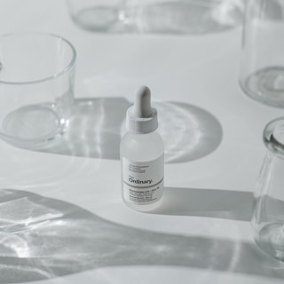 A minimalist arrangement featuring a dropper bottle of skincare product, surrounded by clear, empty glass containers casting soft shadows on a white surface.