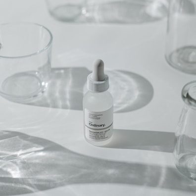 A minimalist arrangement featuring a dropper bottle of skincare product, surrounded by clear, empty glass containers casting soft shadows on a white surface.