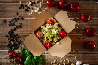 An open cardboard takeout box contains fusilli pasta mixed with pesto sauce, garnished with shredded cheese and fresh cherry tomatoes. Surrounding the box are whole cherry tomatoes, spinach leaves, pumpkin seeds, and scattered cheese on a rustic wooden table.