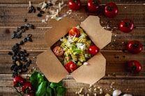 An open cardboard takeout box contains fusilli pasta mixed with pesto sauce, garnished with shredded cheese and fresh cherry tomatoes. Surrounding the box are whole cherry tomatoes, spinach leaves, pumpkin seeds, and scattered cheese on a rustic wooden table.