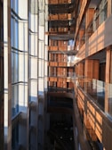 A modern multi-story building interior with a large vertical atrium. Glass windows on the left side allow sunlight to stream in, creating a pattern of light and shadows on the opposite wooden walls and balconies. The building features sleek surfaces, clean lines, and a mix of natural wood and glass materials.