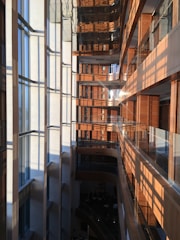 A modern multi-story building interior with a large vertical atrium. Glass windows on the left side allow sunlight to stream in, creating a pattern of light and shadows on the opposite wooden walls and balconies. The building features sleek surfaces, clean lines, and a mix of natural wood and glass materials.